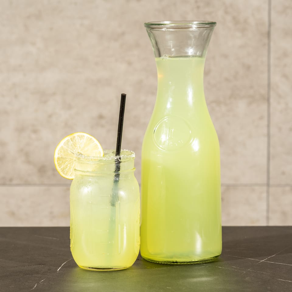 A glass carafe and jar of pale yellow lemonade sit on a dark table, with a lemon slice and black straw.