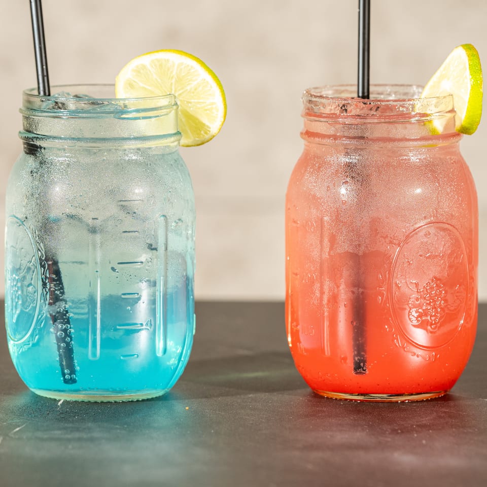 Blue and pink iced drinks in mason jars with black straws and lime wedges on a tabletop.