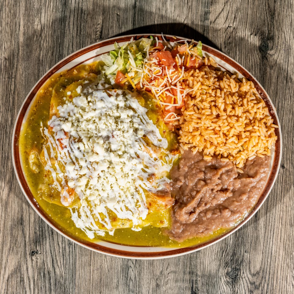 Mexican platter with green enchiladas topped with cream and cheese, Mexican rice, refried beans, and side salad on a wooden table.