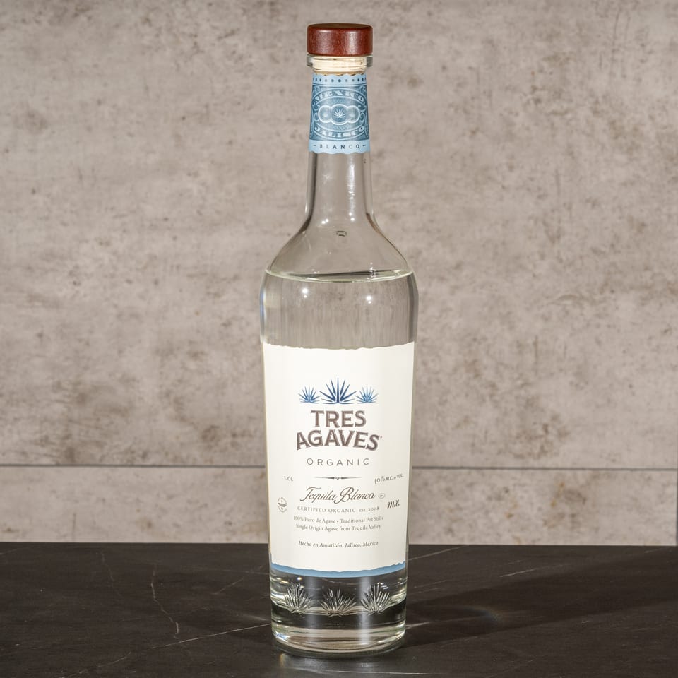 Bottle of Tres Agaves Organic Tequila Blanco displayed on a black countertop against a textured gray tile wall.