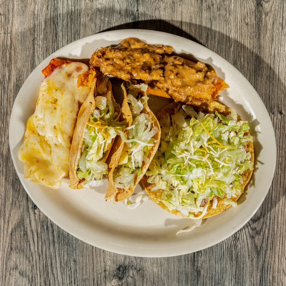 Plate of two crispy lettuce-filled tacos, a cheesy enchilada, and a saucy fried dish on a wooden table.