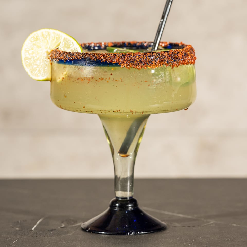 Green margarita in a stemmed glass with a chili-salt rim, lime garnish, and straw on a restaurant table.