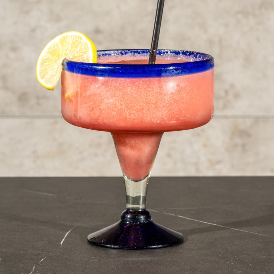 Pink frozen cocktail in a blue-rimmed margarita glass, garnished with a lime wedge and served with a straw.