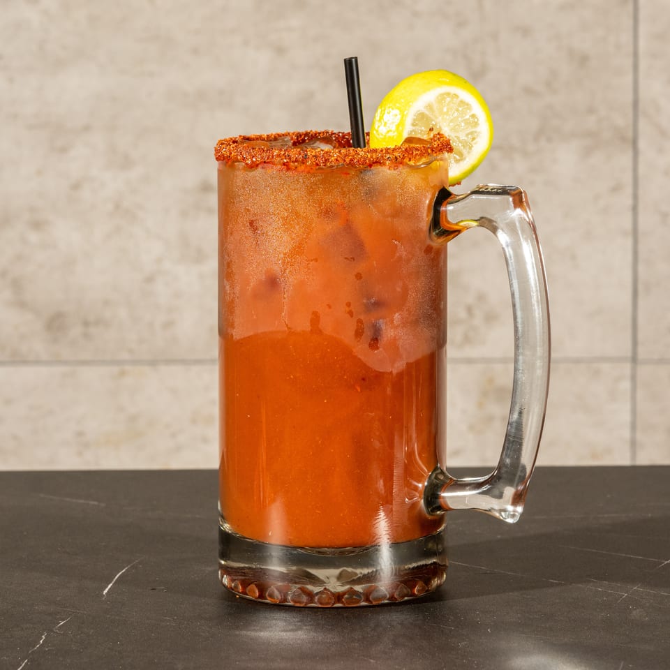 Bloody Mary cocktail in a glass mug with a chili-salt rim, lemon garnish, straw, and ice.