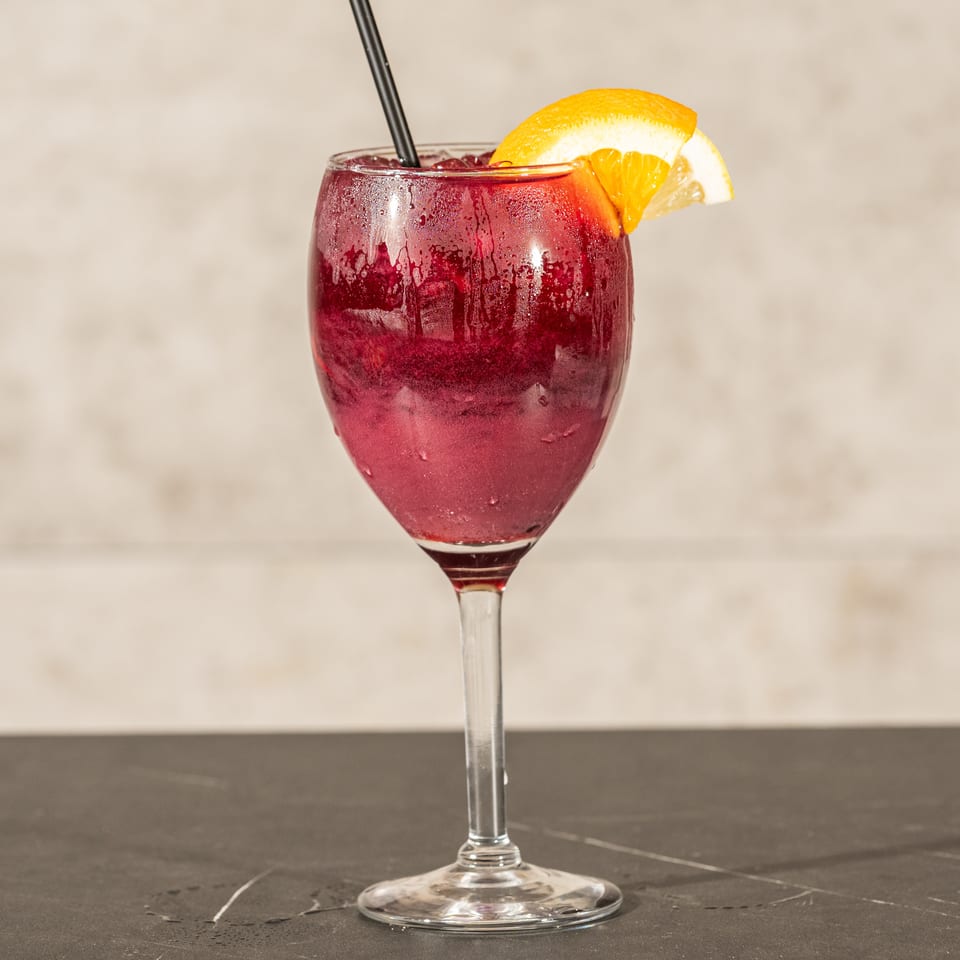Chilled deep-red cocktail in a stemmed glass, garnished with orange wedges and served with a black straw.