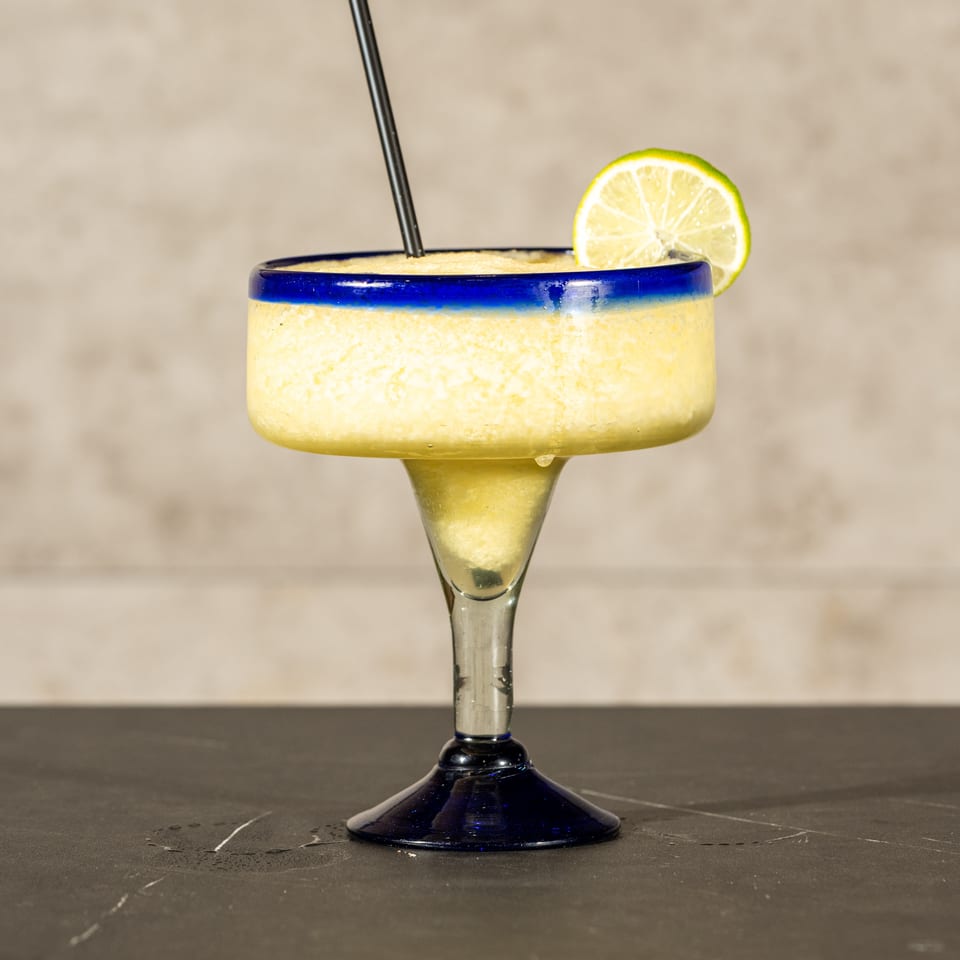 Frosty yellow margarita in a blue-rimmed glass, garnished with a lime slice and straw on a dark table.