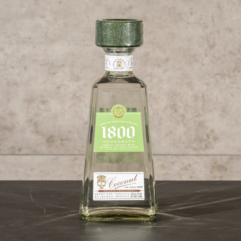 Bottle of 1800 Coconut tequila displayed on a dark tabletop against a textured beige wall.