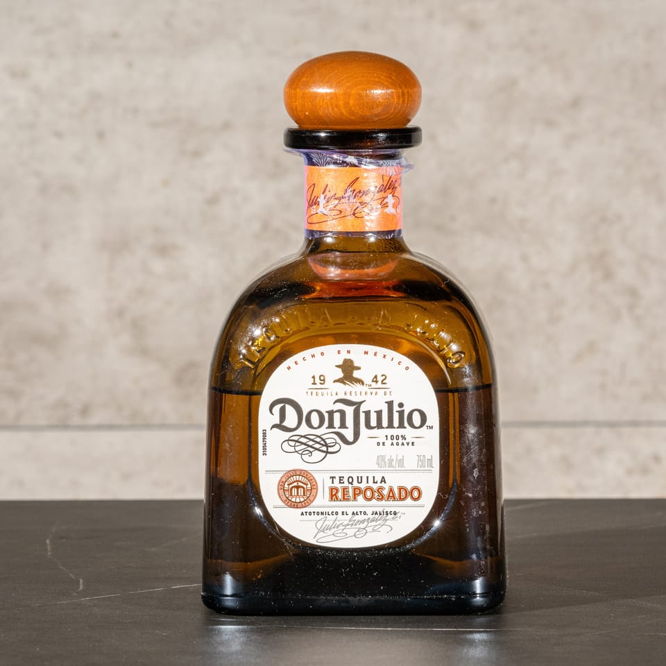 Don Julio Reposado tequila bottle with amber liquid, wooden stopper, and white label displayed on a dark countertop.