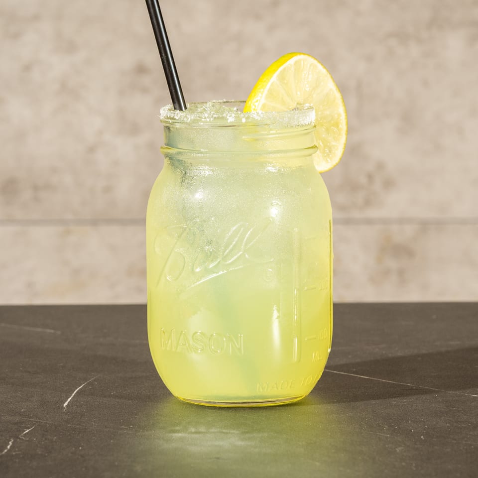 Pale yellow lime drink served in a mason jar with a salted rim, lime wedge, and black straw.