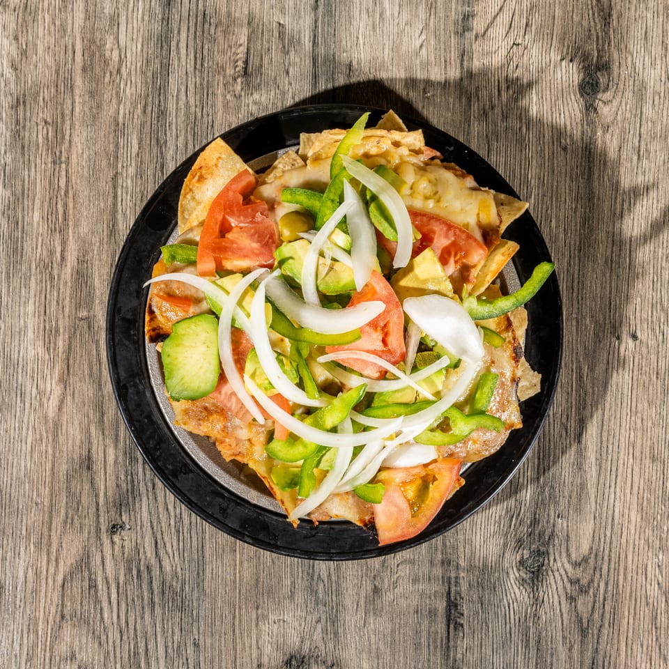 Loaded nachos topped with melted cheese, tomatoes, avocado, green peppers, and sliced onions on a black plate.