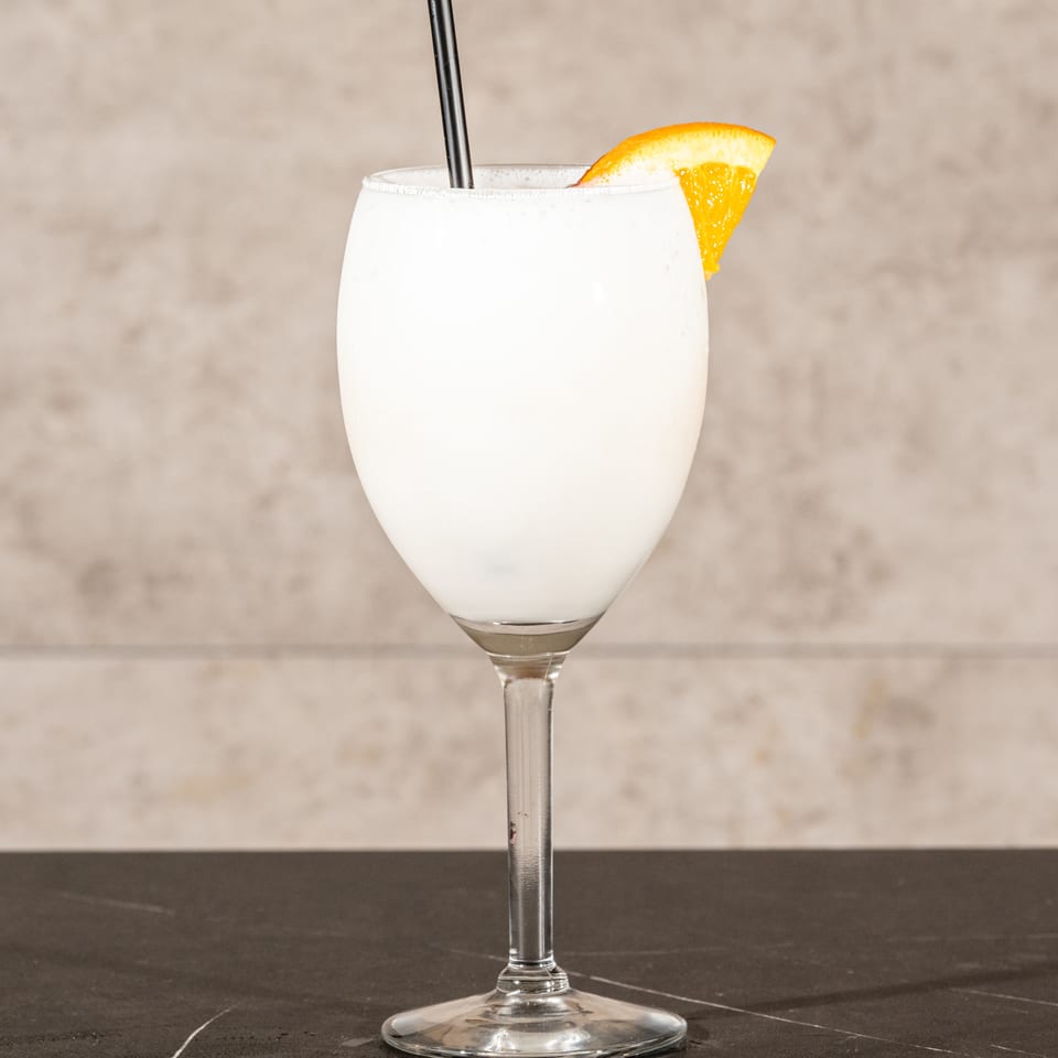 Creamy white cocktail in a stemmed glass, garnished with an orange wedge and served with a black straw.