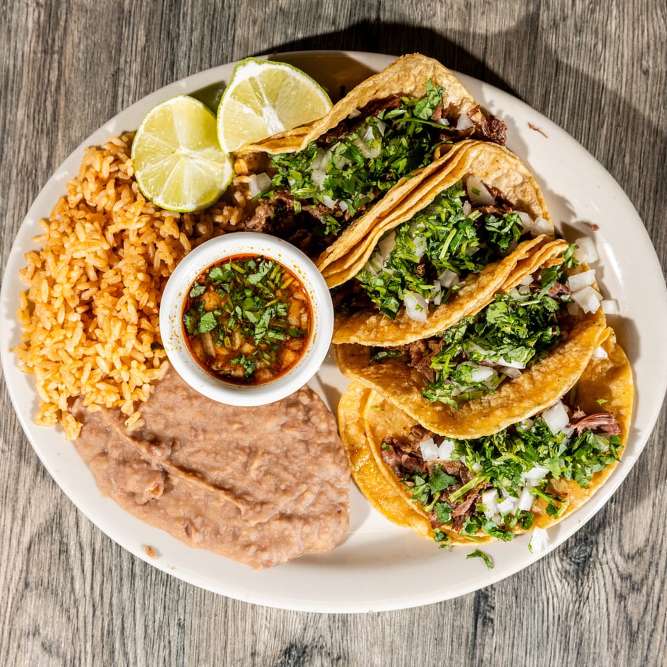 Plate of Mexican tacos topped with cilantro and onions, served with rice, refried beans, lime wedges, and salsa.